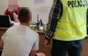 policjant przesłuchuje siedzącego przy biurku zatrzymanego mężczyznę