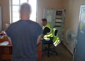 policjant siedzący przy biurku sporządza dokumentację w związku z zatrzymaniem mężczyzny