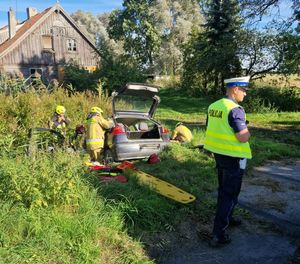 policjant stoi obok miejsca wypadku widać samochód w rowie i stażaków