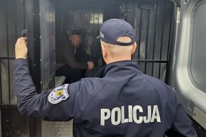 policjant stoi przed otwartym radiowozem w środku siedzi zatrzymany mężczyzna