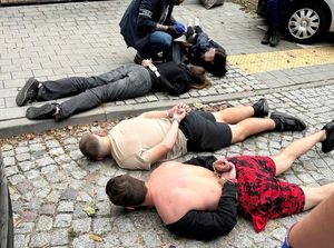 osoby zatrzymane leża na chodniku, mają założone kajdanki. Przy jednym z nich klęka kuca policjant