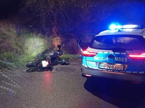 motocykl leżący przy jezdni obok zaparkowany radiowóz z włączonymi sygnałami świetlno dźwiękowymi