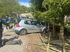 na zdjęciu widać samochód sprawców, którym wjechali w policyjny radiowóz i ogrodzenia. Na zdjęciu stoi policjant i na ziemi leży podejrzany, który na ręce trzymane z tyłu ma założone kajdanki. W tle inni policjanci i radiowóz.